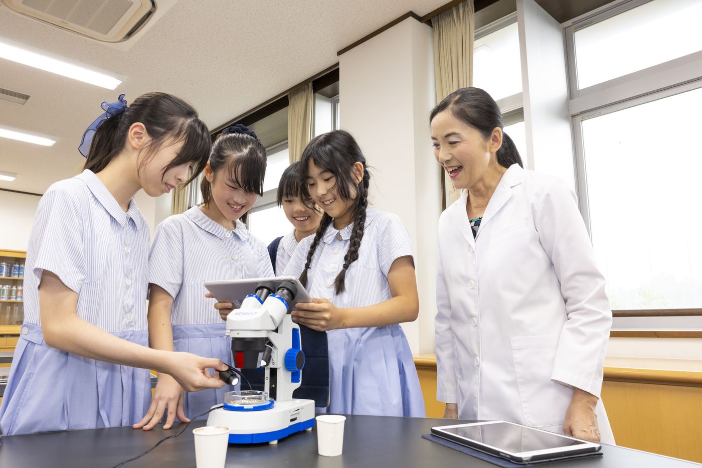 理科の授業で、白衣の先生が顕微鏡をのぞく児童たちのすぐ横で笑顔を見せている写真